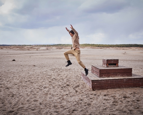 Na zdjęciu mężczyzna skacze z drewnianej konstrukcji. Ręce unosi wysoko do góry. Ma na sobie beżowy golf i spodnie. Wokół piaski pustyni oraz otaczający ją las.